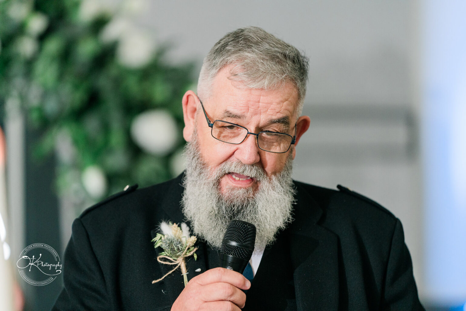 An elderly man with a grey beard and glasses speaks into a microphone, wearing a dark jacket and a boutonniere, with greenery in the background.