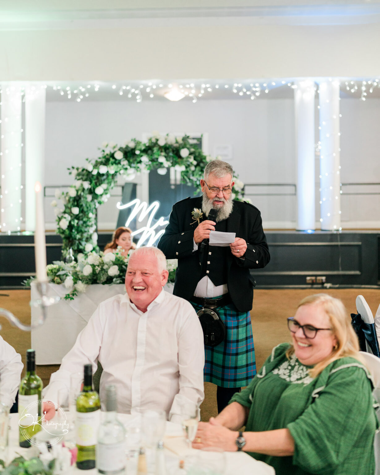 An elderly man in a kilt gives a speech at a wedding reception, while guests smile and laugh at their table. The room is decorated with fairy lights and a floral arch.