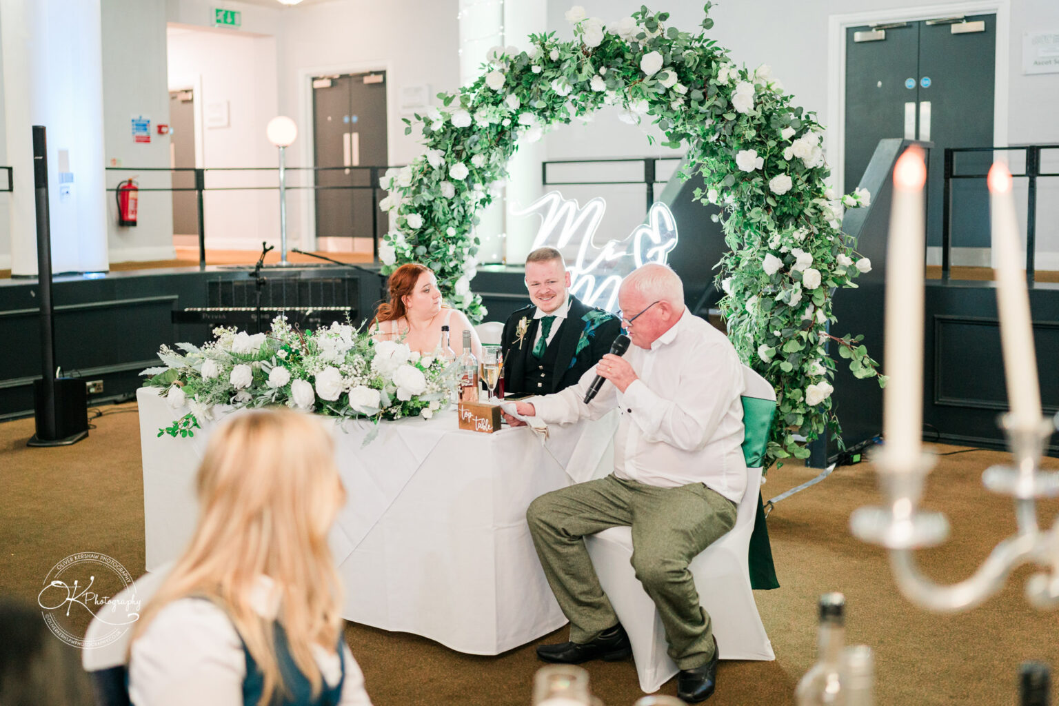 A couple sitting at a decorated table with floral arrangements and a neon "Mr & Mrs" sign, while an elderly man gives a speech using a microphone.
