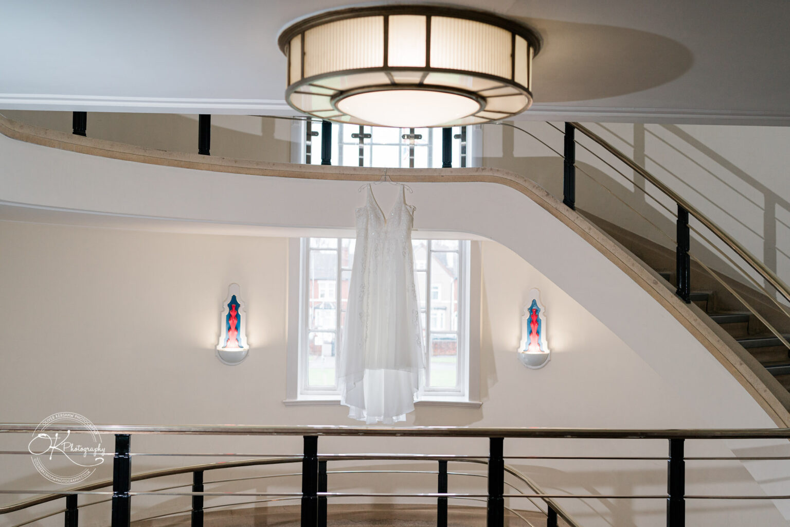 A white wedding dress hanging inside a modern conference centre with a curved staircase, railings, and wall sconces.