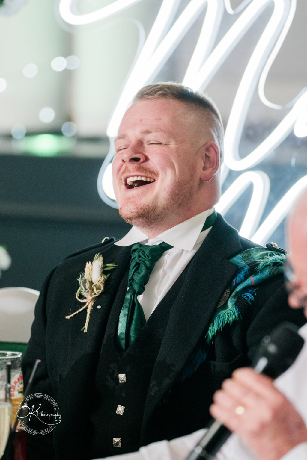 A man in a traditional Scottish outfit laughing, with a boutonniere on his jacket, at a formal event.