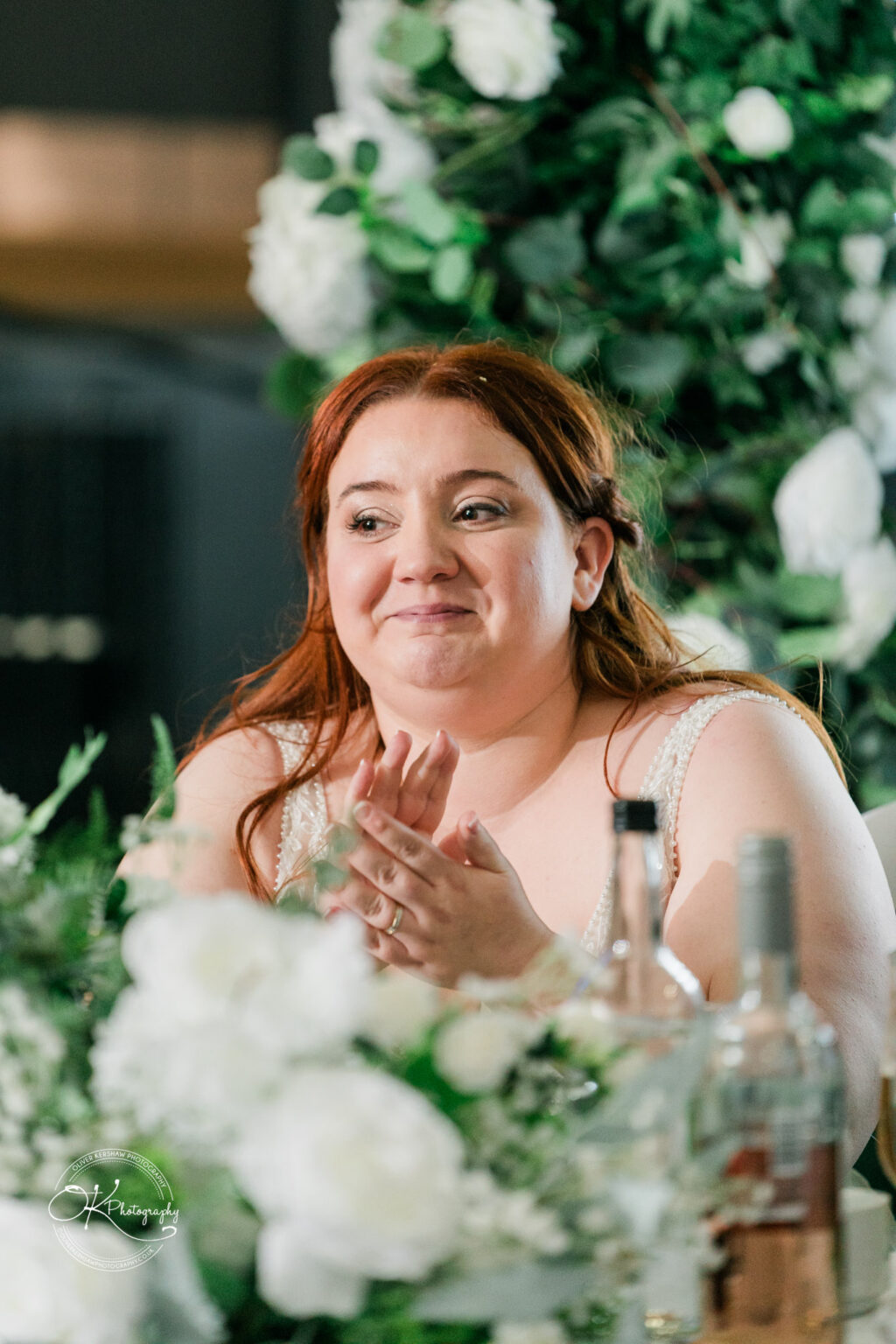 A woman with red hair sits at a table, clapping and smiling, surrounded by white flowers and greenery.