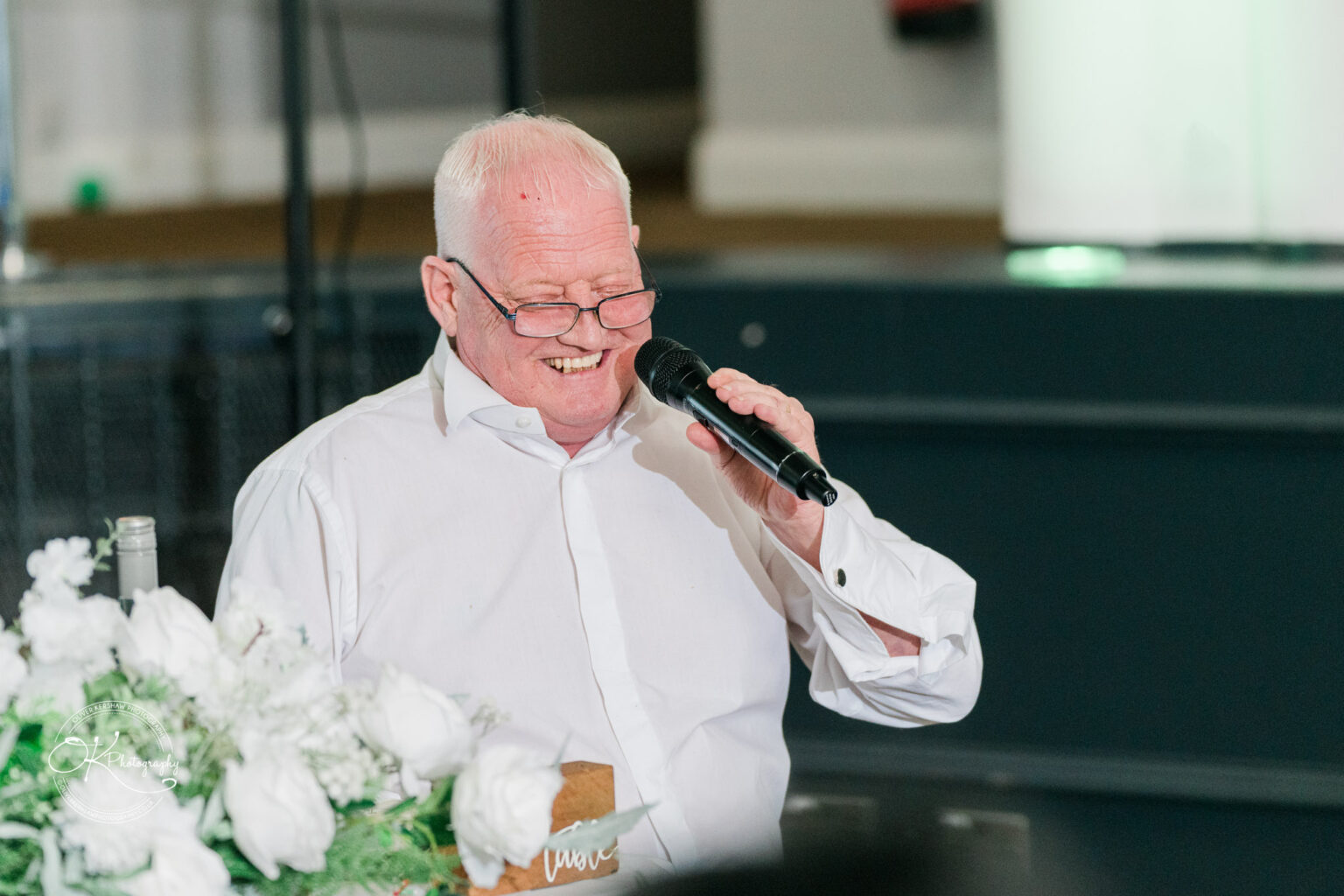 An elderly man in a white shirt speaking into a microphone at a conference, with a bouquet of white flowers in the foreground.