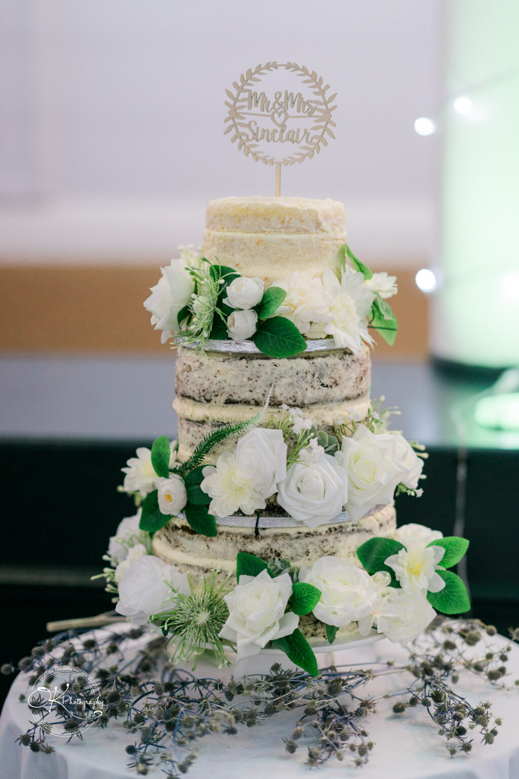 A multi-tier naked wedding cake decorated with white flowers and greenery, topped with a wooden sign that reads "Mr & Mrs Sinclair" on a table with a white tablecloth at an event venue.
