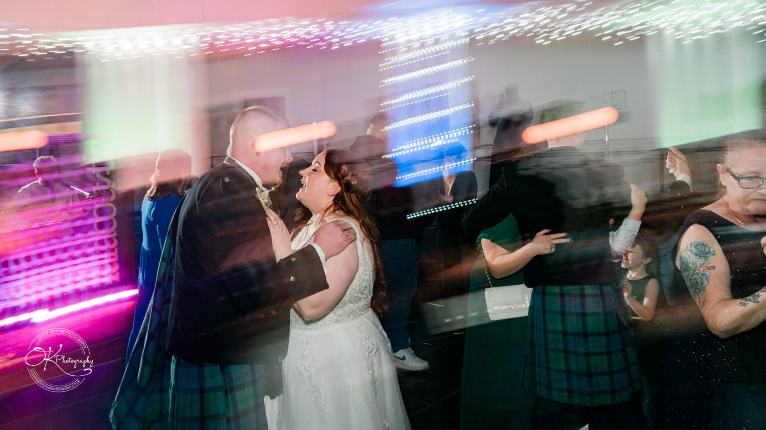 A bride and groom dance at a wedding reception with other guests dancing in the background under colourful lights.