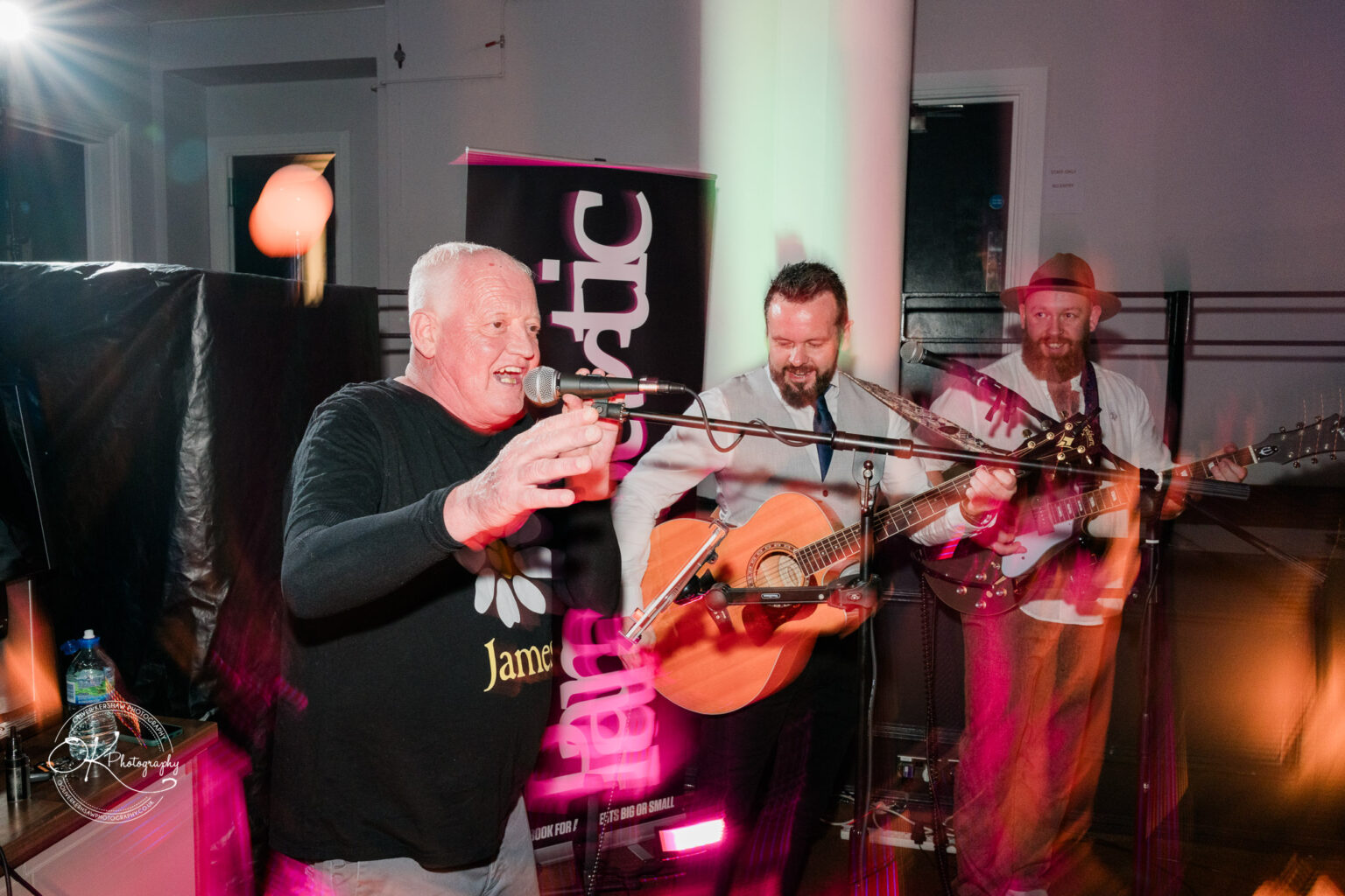 A man singing into a microphone with two other musicians playing guitars on a stage, with colourful lights and a banner in the background.