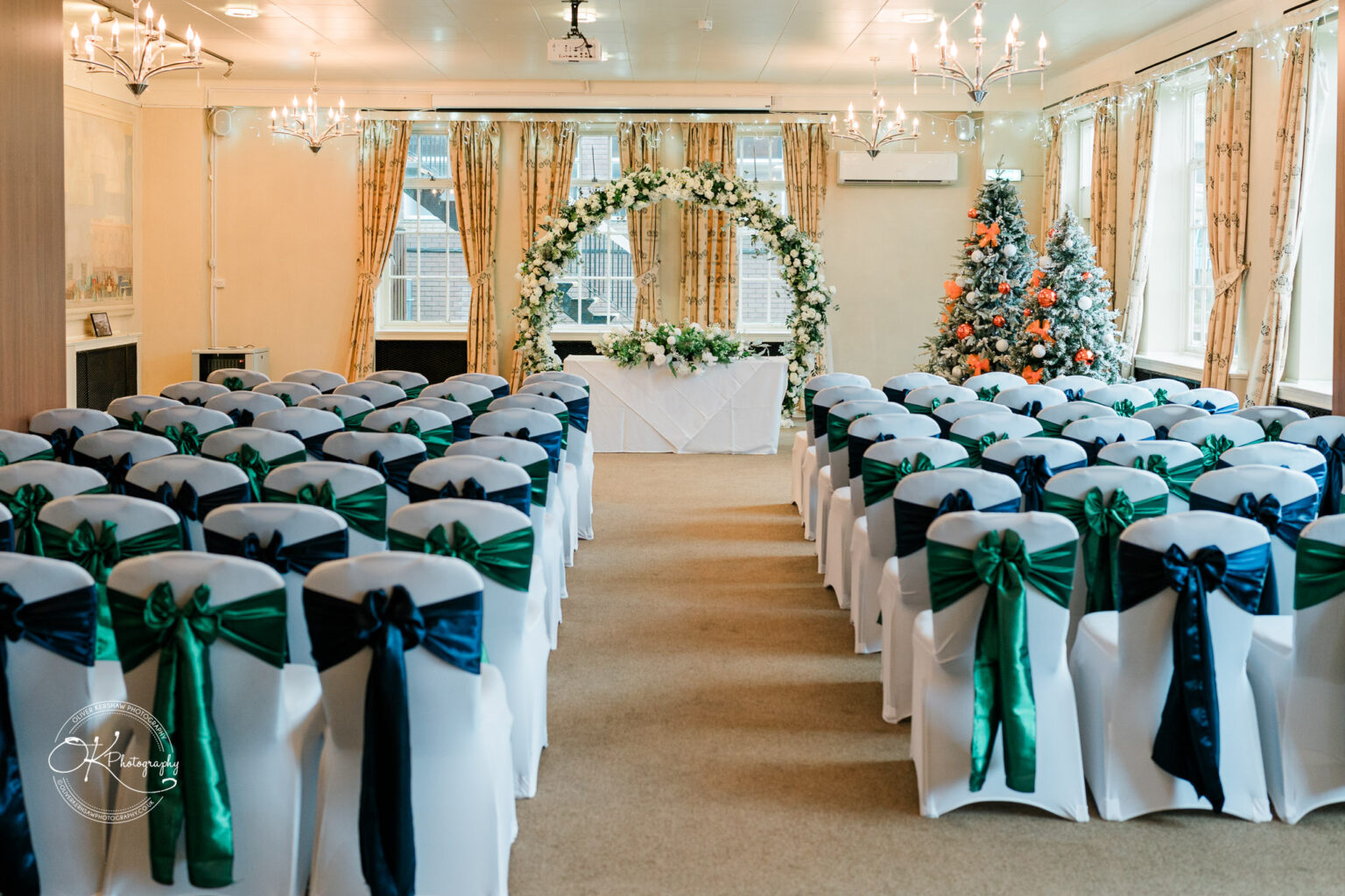 A decorated conference room with rows of white chairs adorned with blue and green bows, a floral arch at the front, and a Christmas tree on the right side.