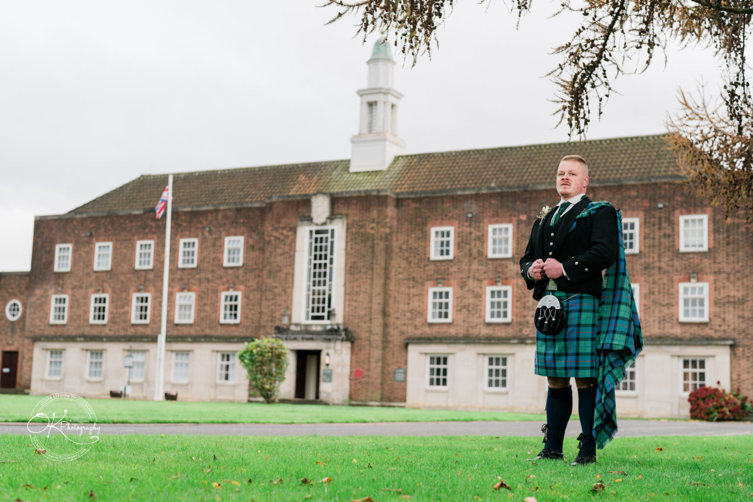 A man in traditional Scottish attire stands on a grassy lawn in front of the Derby Conference Centre building.