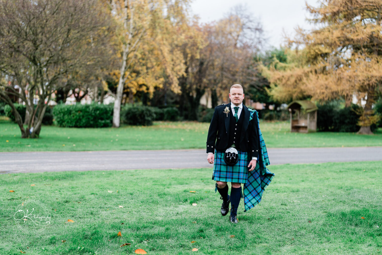 A man in traditional Scottish attire stands on a grass field with autumn trees in the background.