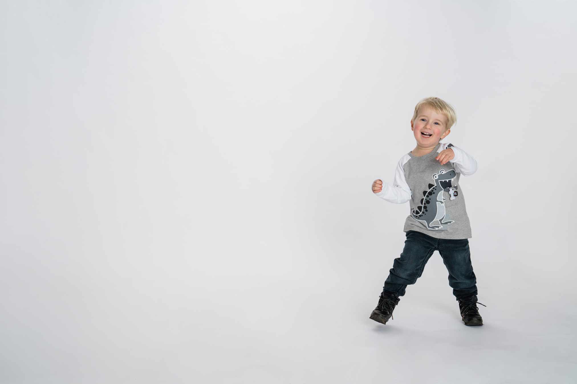 Professional studio photo of a happy blonde toddler dancing in a dinosaur shirt and jeans, set against a white background. Perfect for capturing joyful childhood moments.