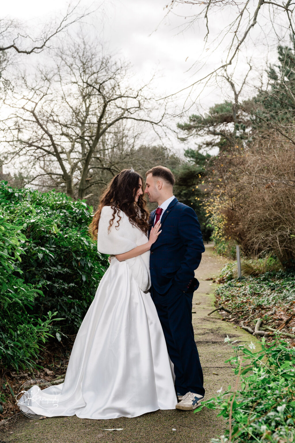 A couple in wedding attire stands close together on a garden pathway, the bride in a white gown and fur stole, and the groom in a dark suit.