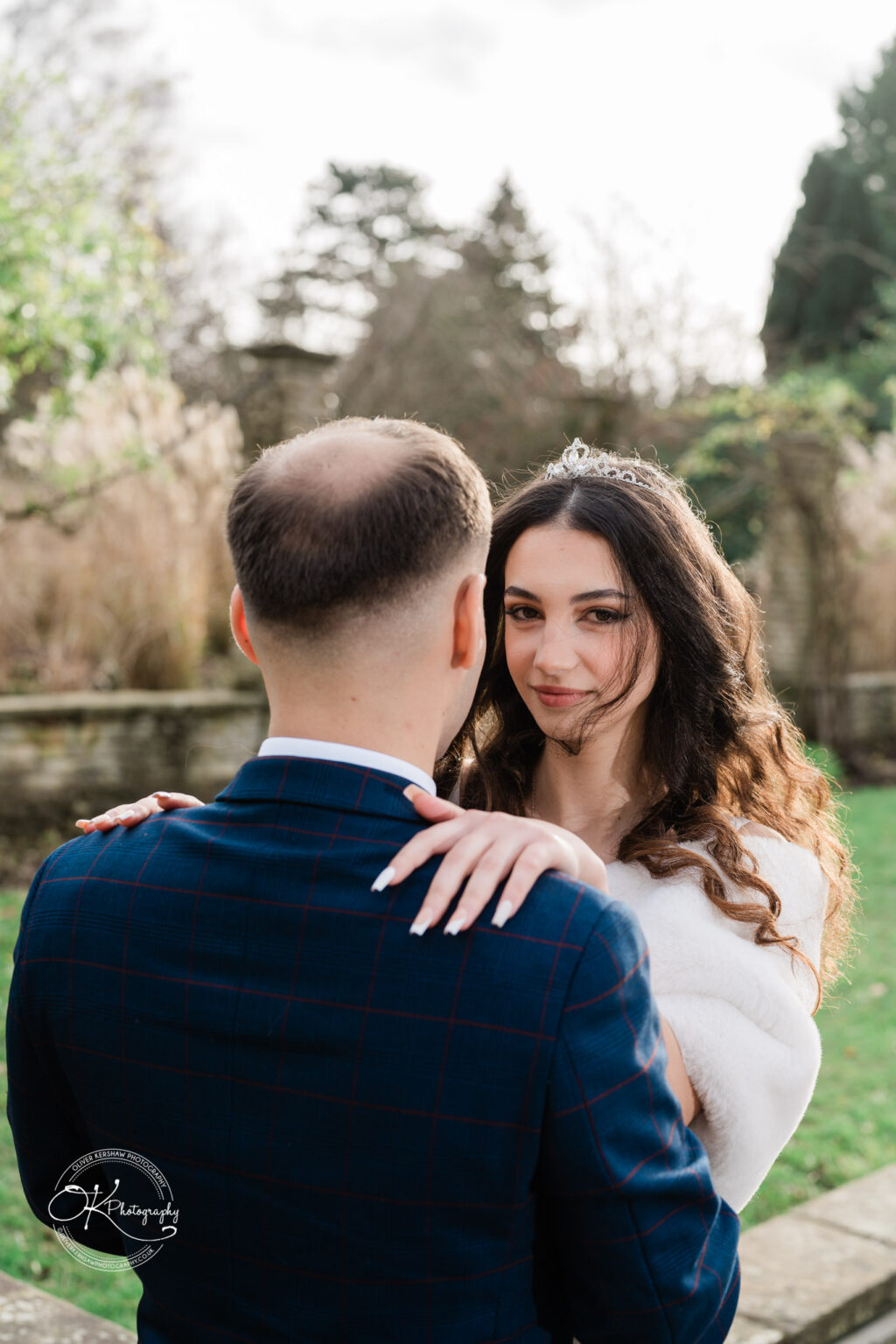 A bride with long, wavy hair and a tiara embraces the groom, who is wearing a dark blue suit, in an outdoor setting.