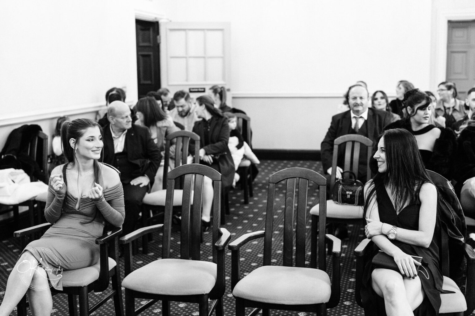 A group of formally dressed people seated and chatting in a room with wooden chairs, two women in the foreground smiling at each other.