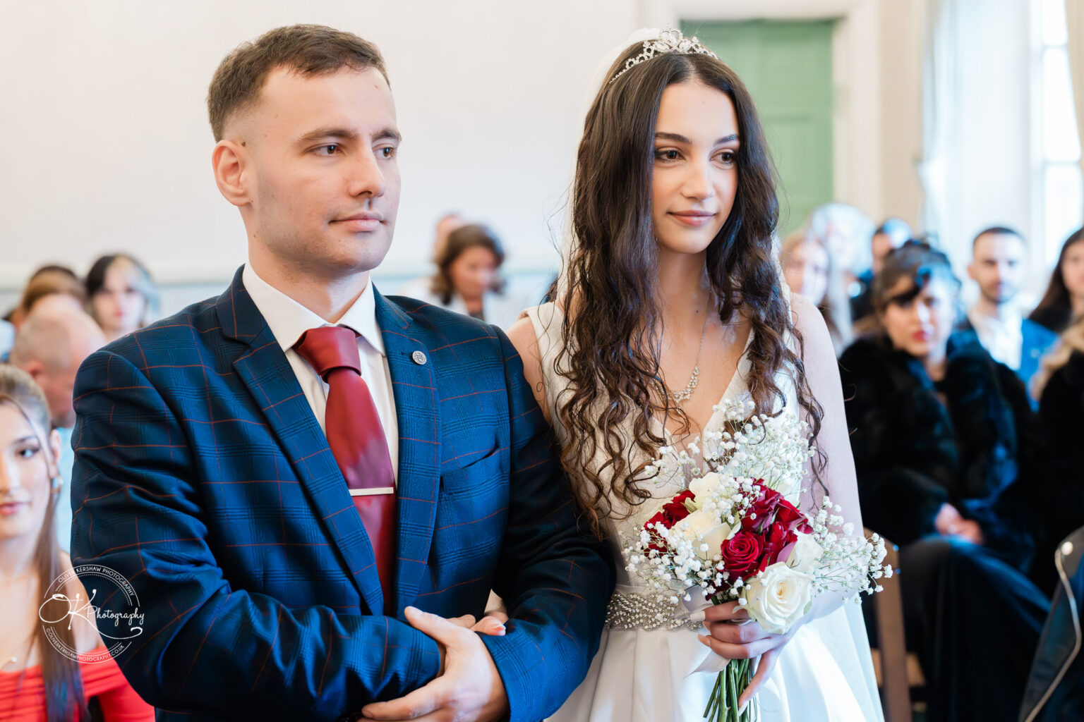 Bride and groom standing together during their wedding ceremony, with the bride holding a bouquet of red and white flowers.