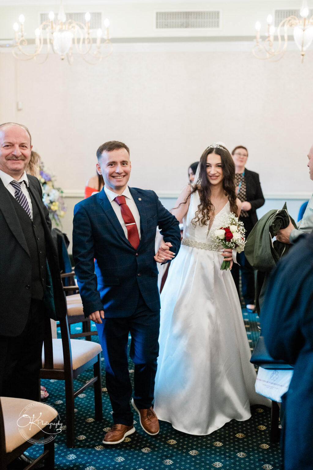 Groom and bride walking down the aisle, smiling and holding hands, with guests in the background.
