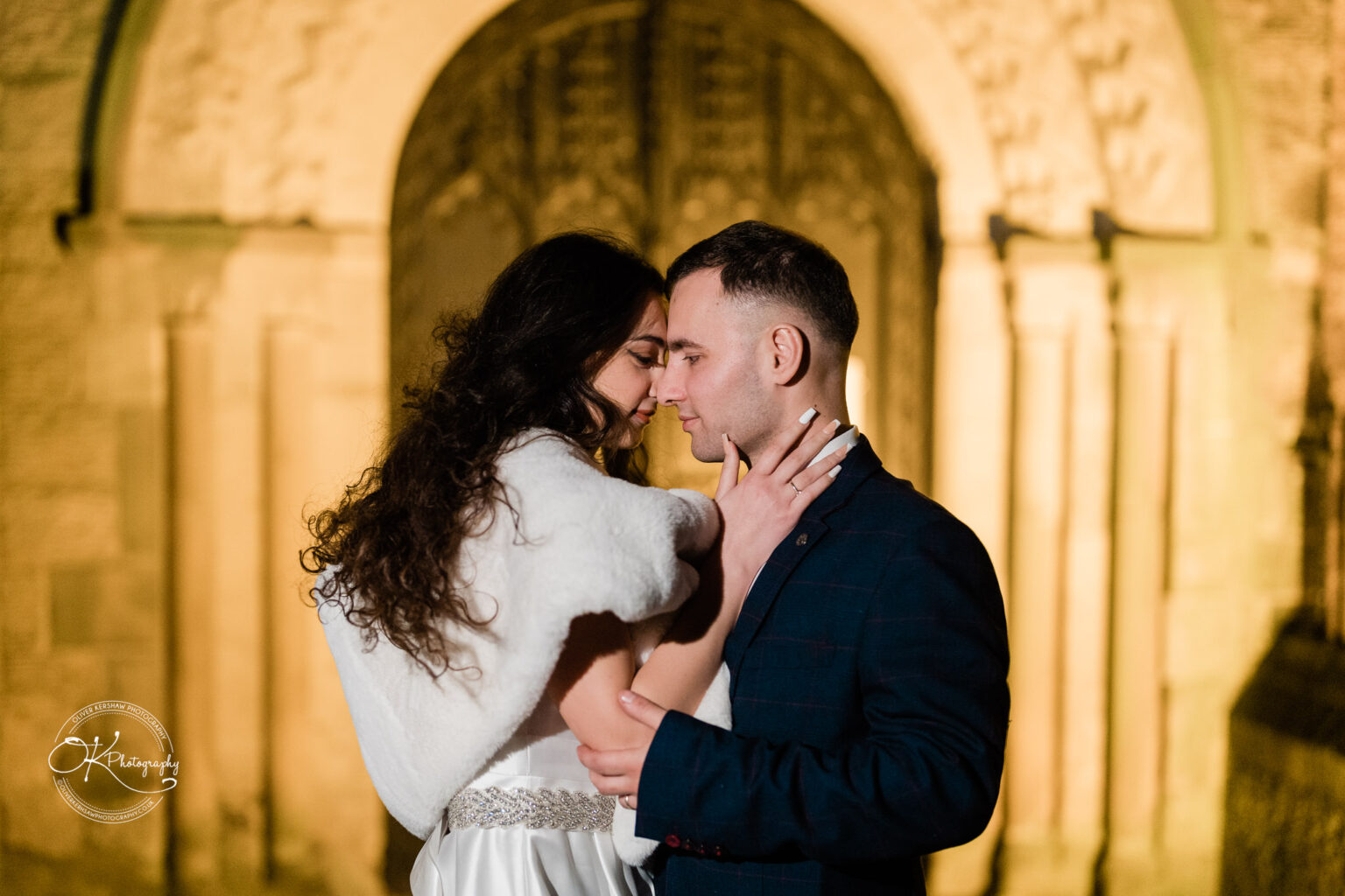 A couple embracing closely in front of an ornate, lit-up archway.