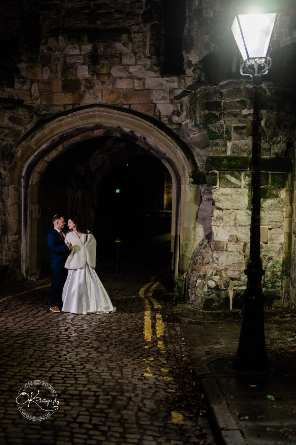 Bride and groom standing under an archway at night, illuminated by an old-fashioned street lamp.