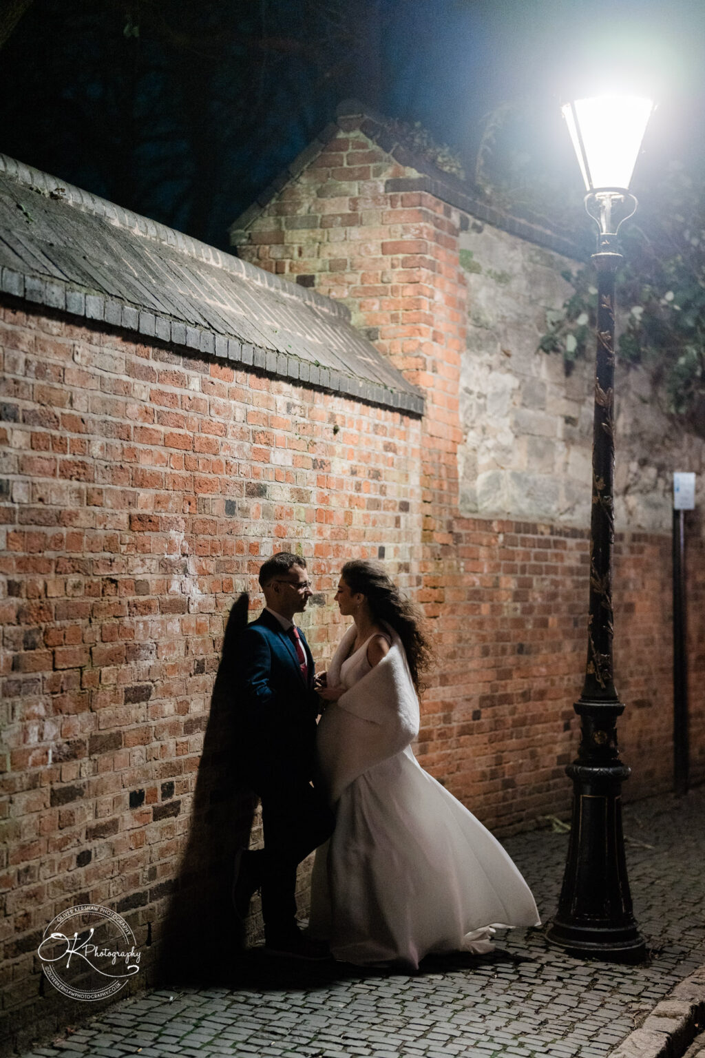 A couple standing close together next to a brick wall at night, illuminated by a street lamp.