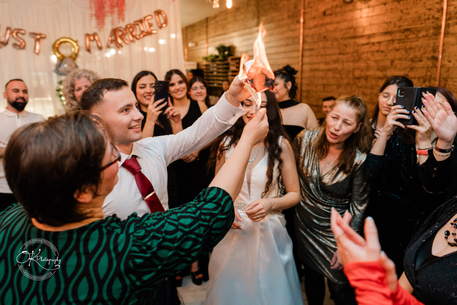 Wedding celebration with guests surrounding a couple, holding a flaming object, under "Just Married" balloons.