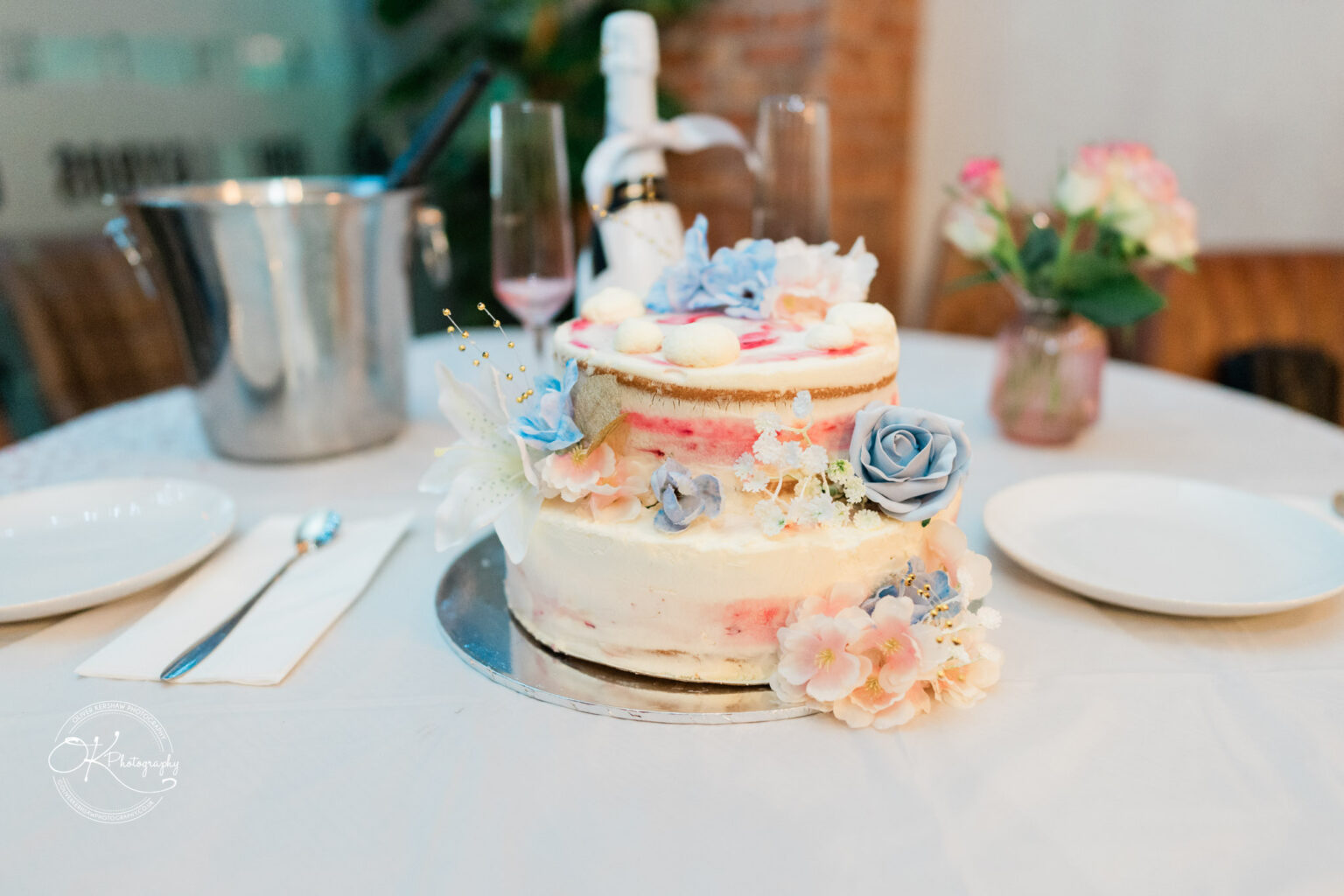 Two-tiered cake decorated with blue, pink, and white flowers on a white table with plates, cutlery, a bottle of wine, and a vase of flowers.
