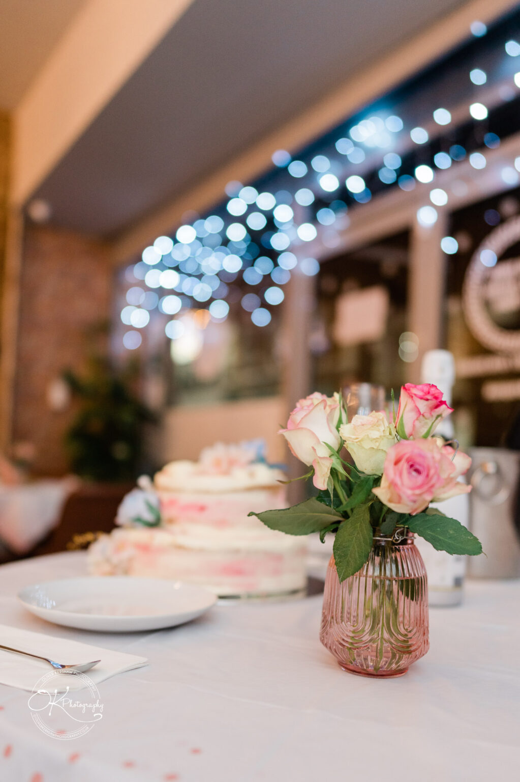 Bouquet of pink and white roses in a glass vase on a table with a blurred background of a cake and twinkling lights.