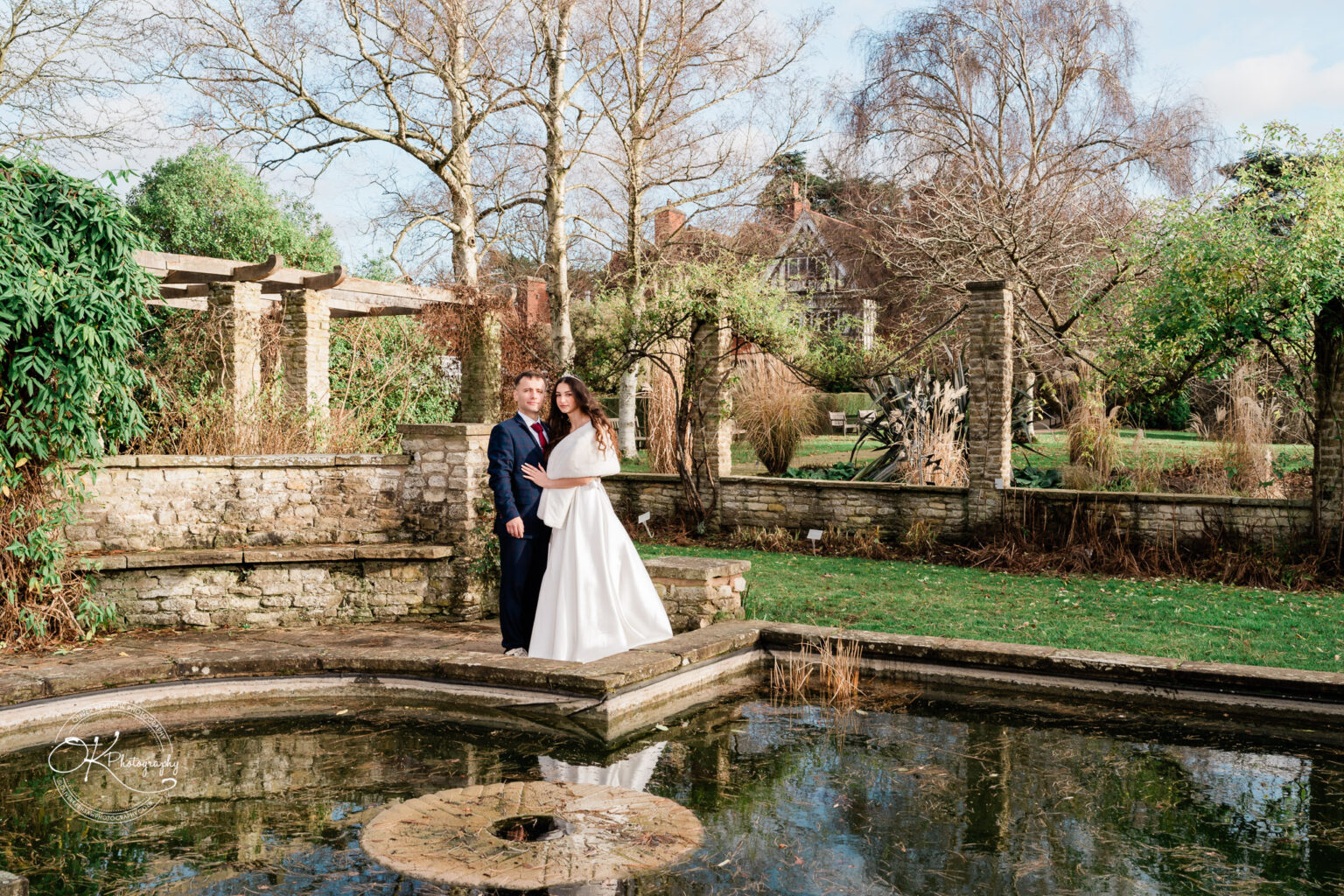 A couple in formal attire posing beside an outdoor pond in a garden with stone structures and trees in the background.