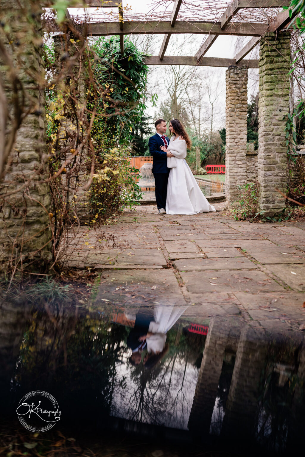 Bride and groom embrace under a wooden trellis in a garden with their reflection visible in a puddle on the ground.