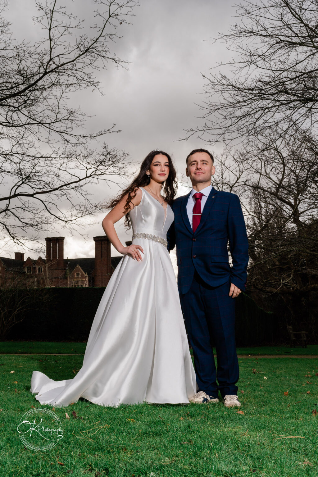 Bride and groom standing on a lawn with bare trees and a building in the background, under a cloudy sky.