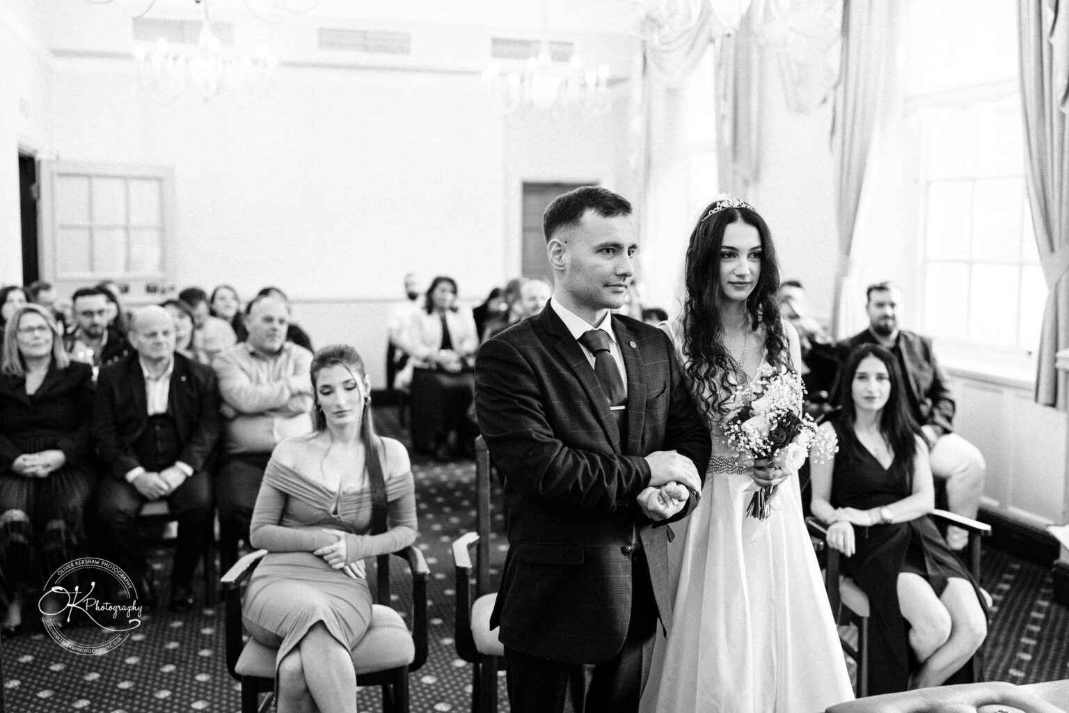 Bride and groom standing in front of seated guests during a wedding ceremony in a formal room.