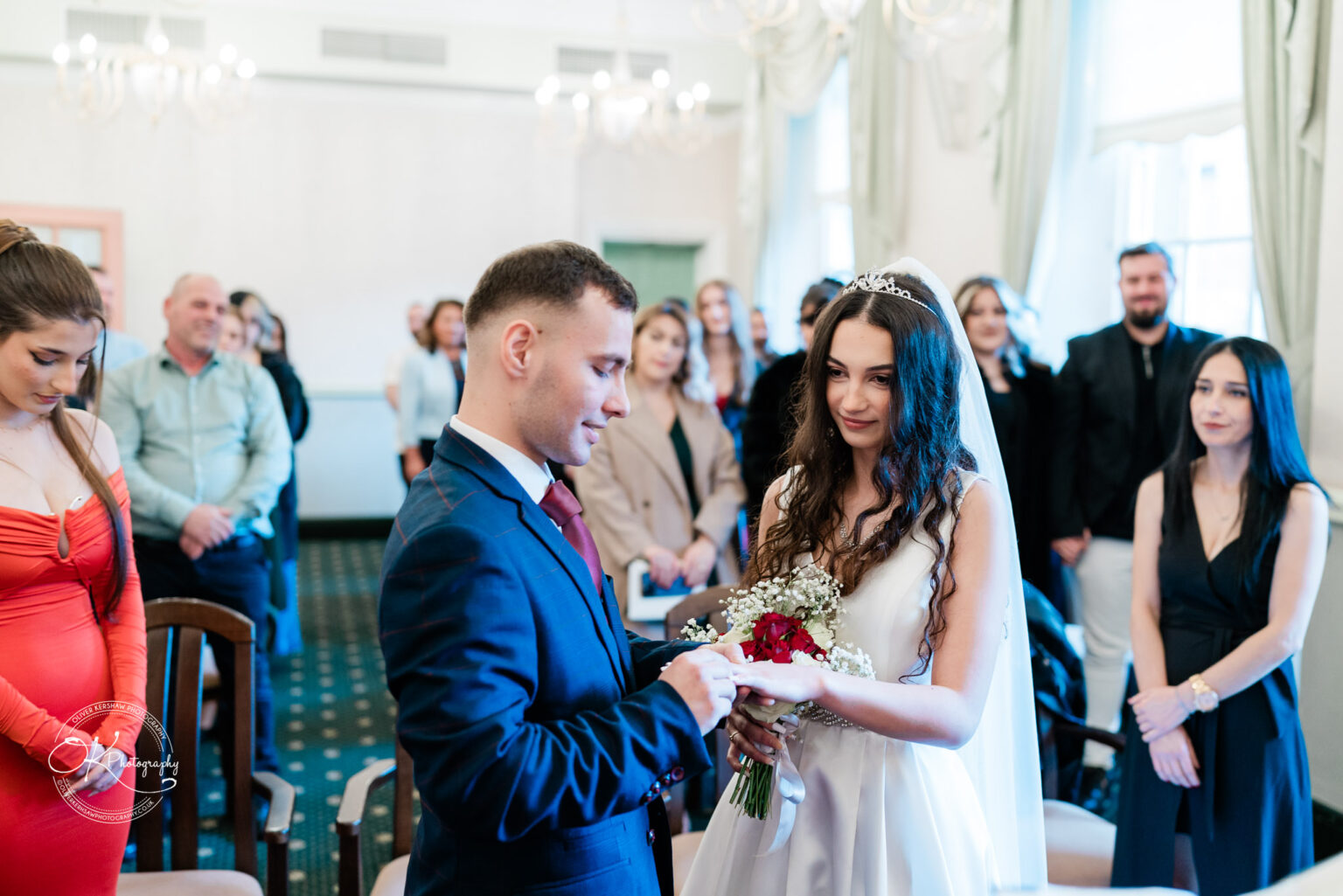A bride and groom exchange rings during a wedding ceremony, surrounded by family and friends in a well-lit room.
