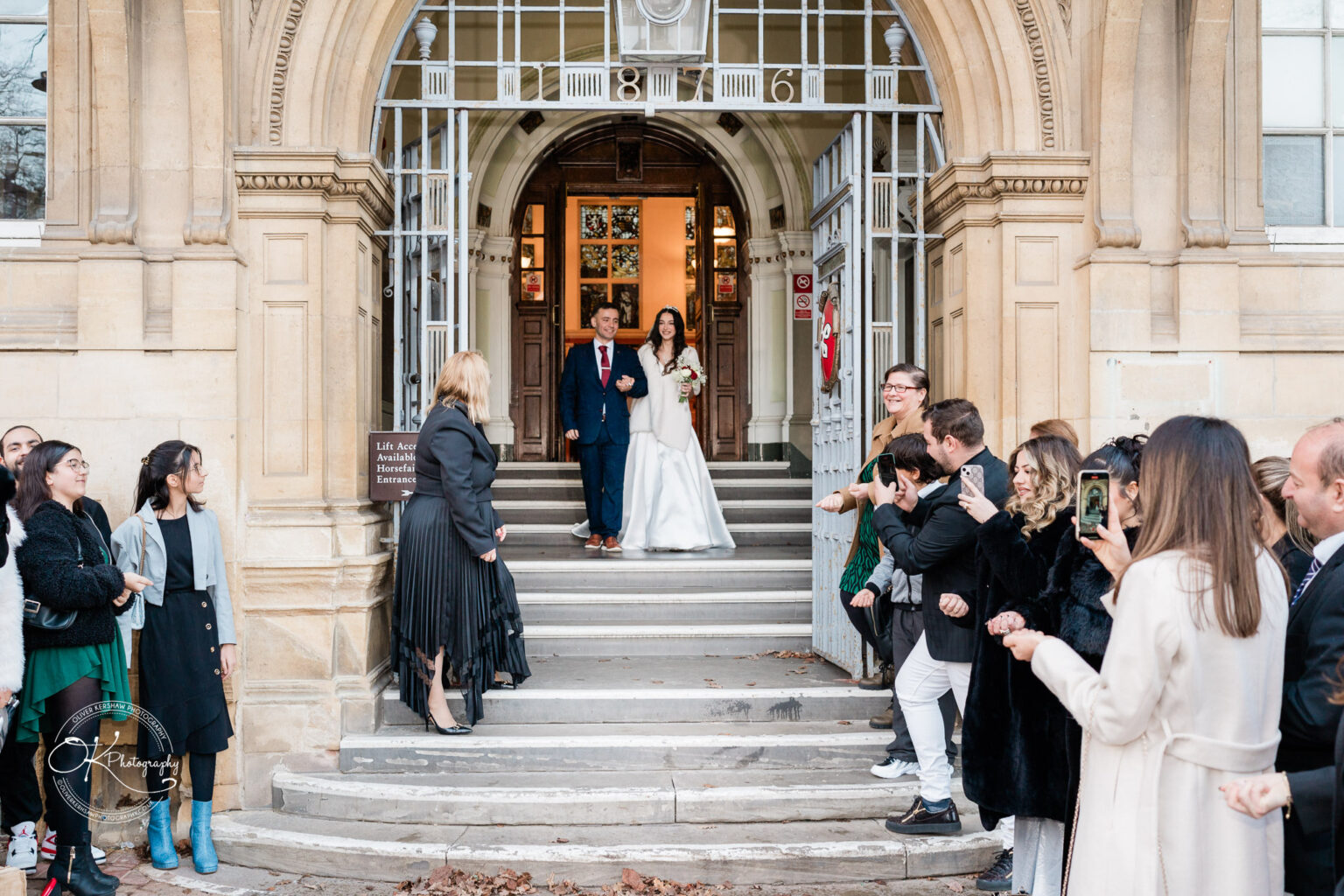 A bride and groom stand arm in arm at the top of a set of stairs, exiting a building under an arch with the date "1876" above the door. Guests are gathered on both sides of the stairs, some taking photos with their phones.
