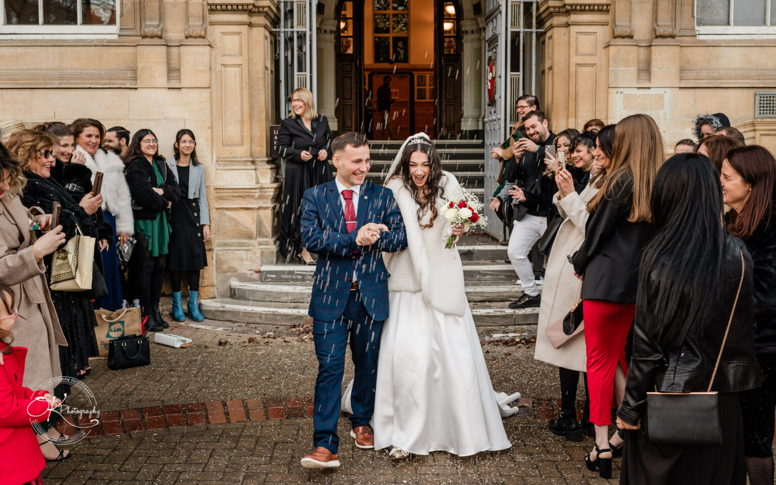 Bride and groom walking down steps outside a building, surrounded by guests taking photos and smiling.