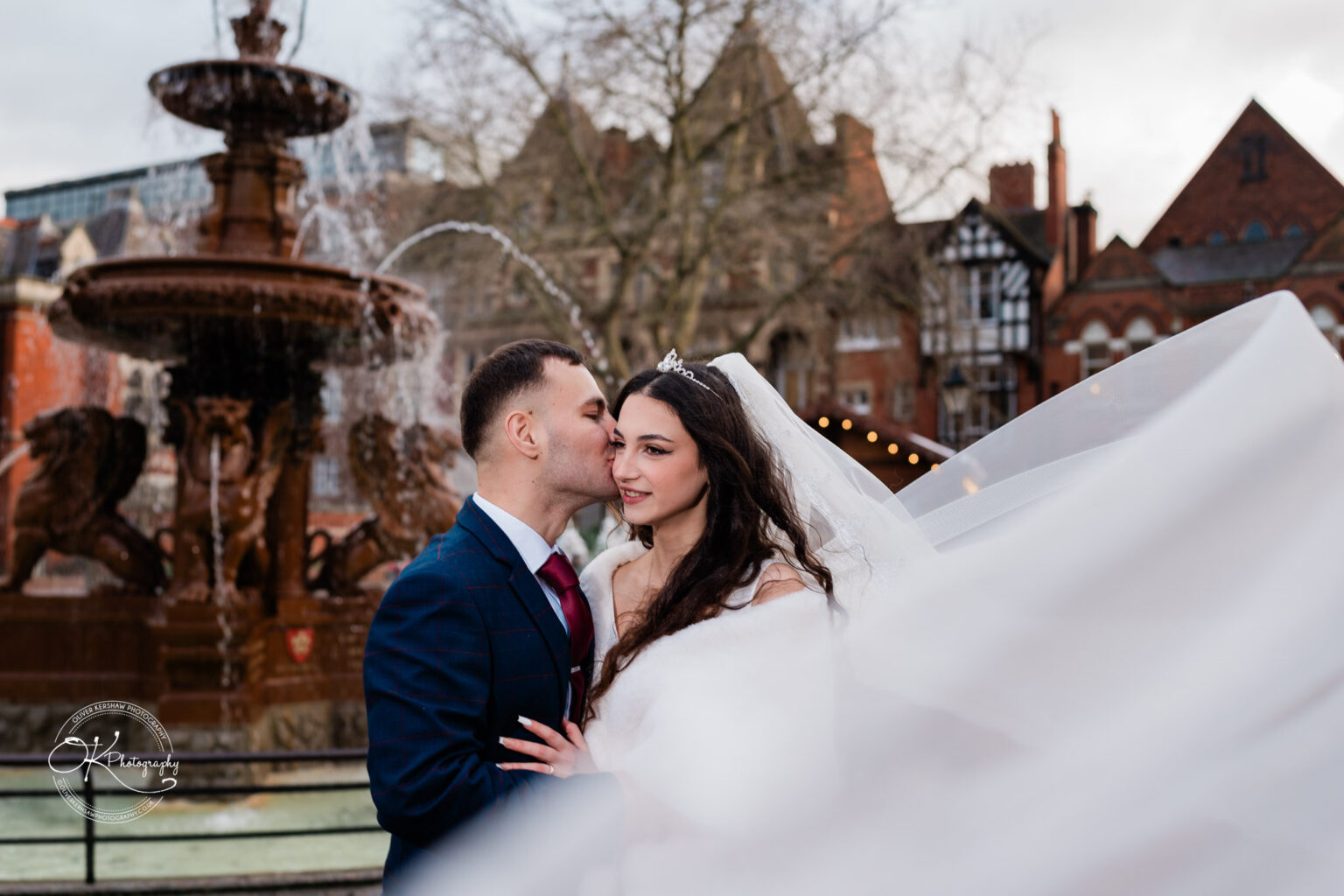Bride and groom in wedding attire embrace in front of a decorative fountain, with historic buildings in the background.