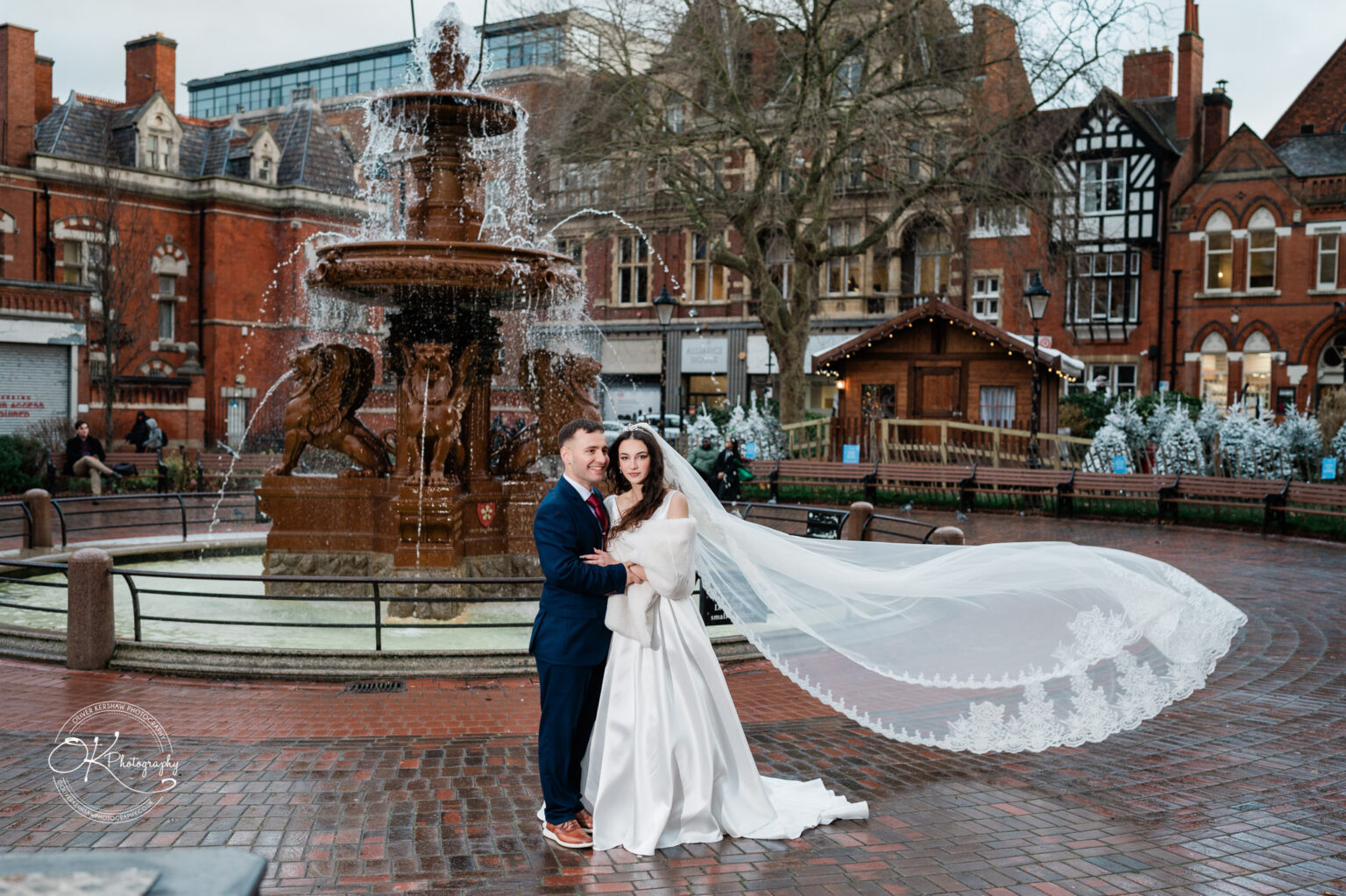 Bride and groom standing in front of a fountain in an urban setting with historic buildings in the background.