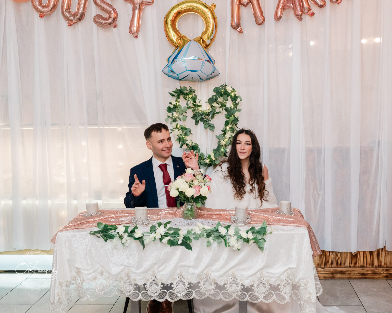 A groom in a navy suit with a red tie and a bride in an off-shoulder white dress sit at a table adorned with flowers and candles. Behind them is a backdrop of white curtains, balloons, and greenery forming a heart shape.