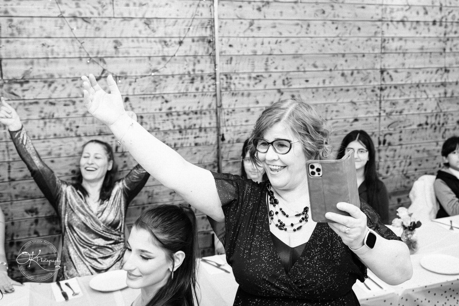 A joyful woman with glasses raises her arm and holds a smartphone, surrounded by smiling people at a celebratory gathering, with a wooden wall in the background.