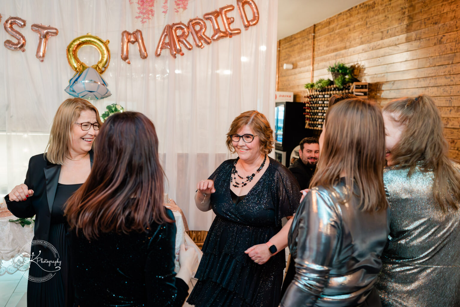 A group of people dancing and laughing at a celebration with rose gold "Just Married" balloons in the background.