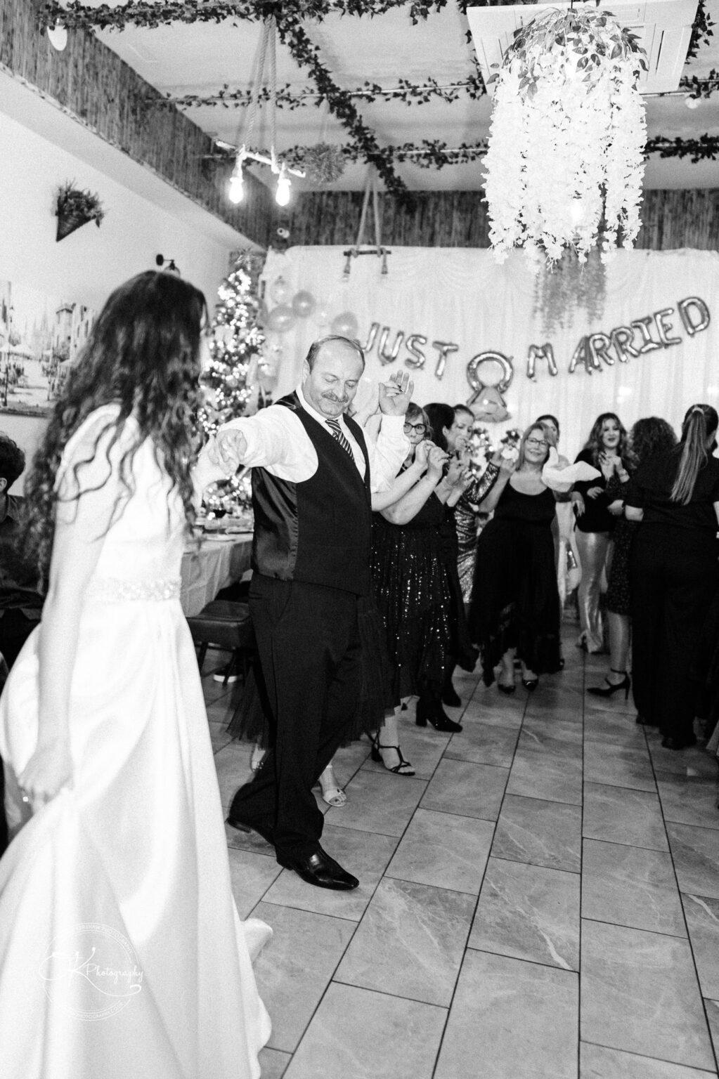Black and white image of a wedding celebration, with people dancing in a line under a "Just Married" banner.
