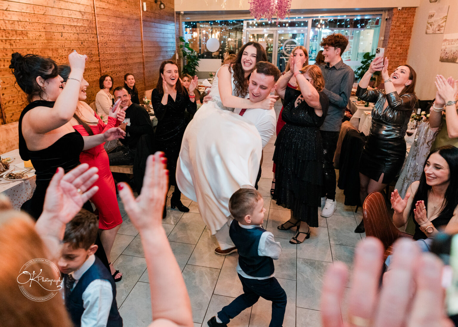 Bride and groom dancing and laughing, surrounded by cheering and clapping guests at a wedding reception.