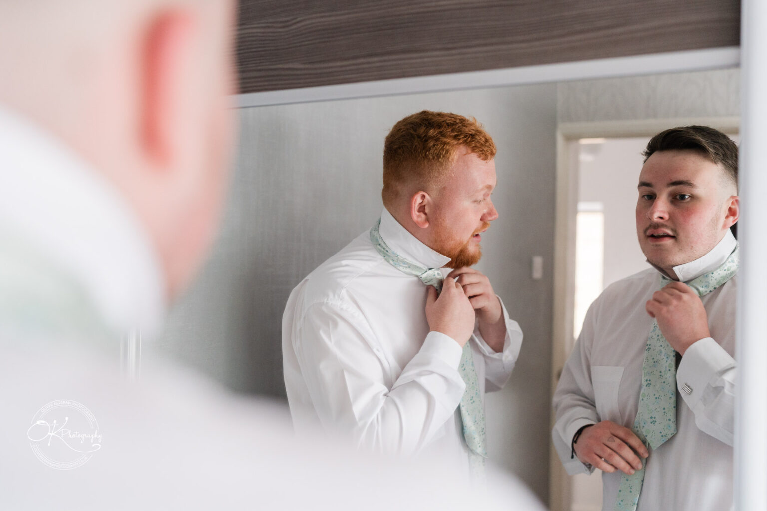 Two men standing in front of a mirror, adjusting their ties, both dressed in white shirts.