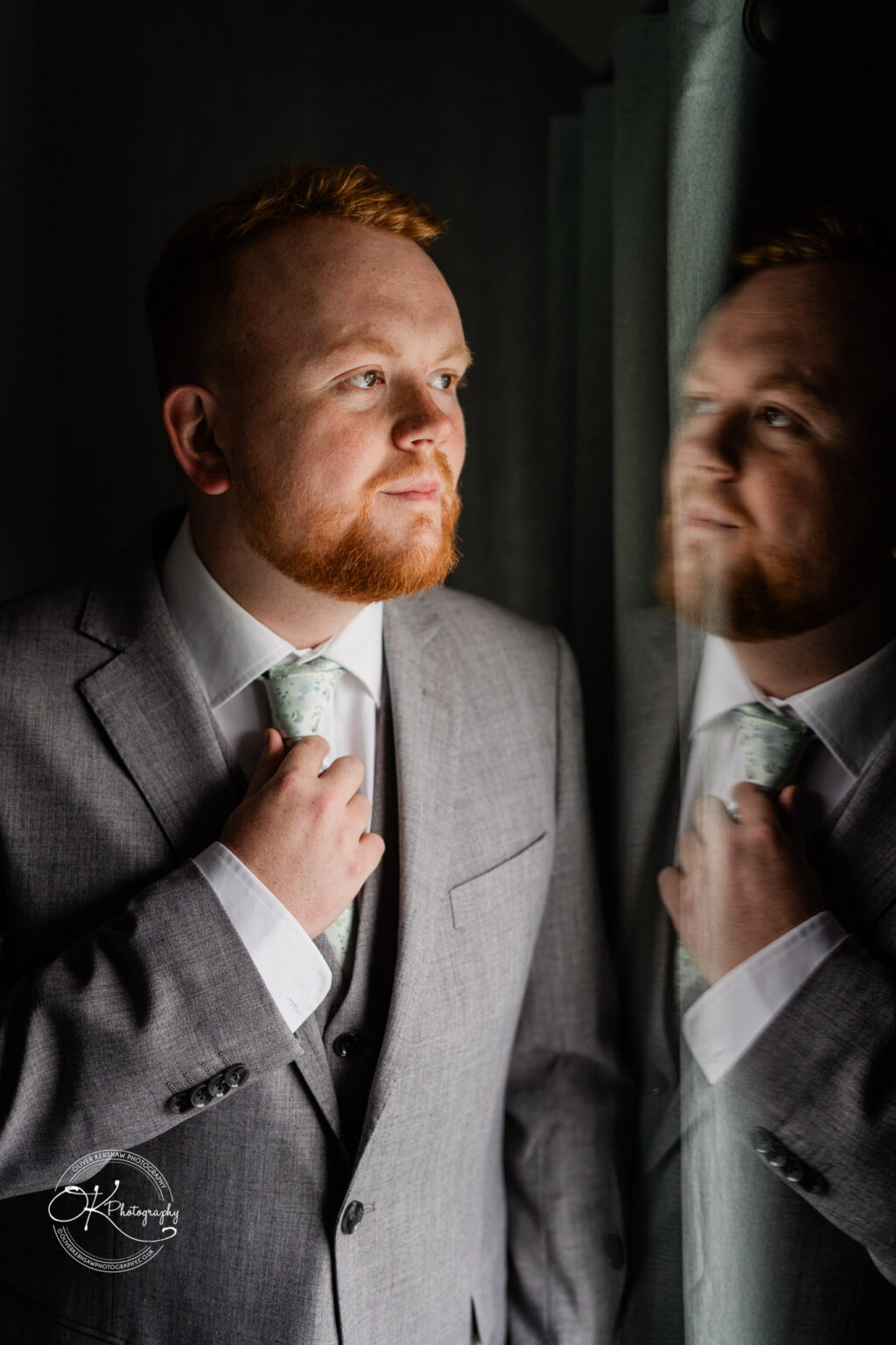 A man with red hair and a beard, wearing a grey suit and adjusting his tie while looking out of a window.