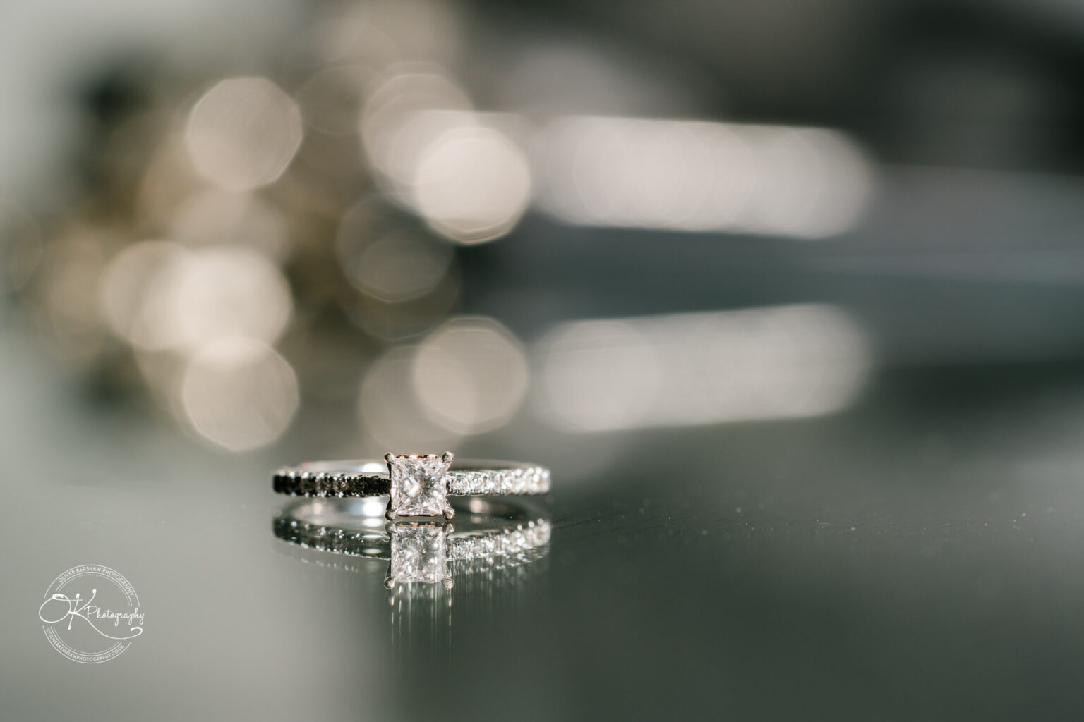 A close-up of an engagement ring with a square-cut diamond, placed on a reflective surface with a blurred background.
