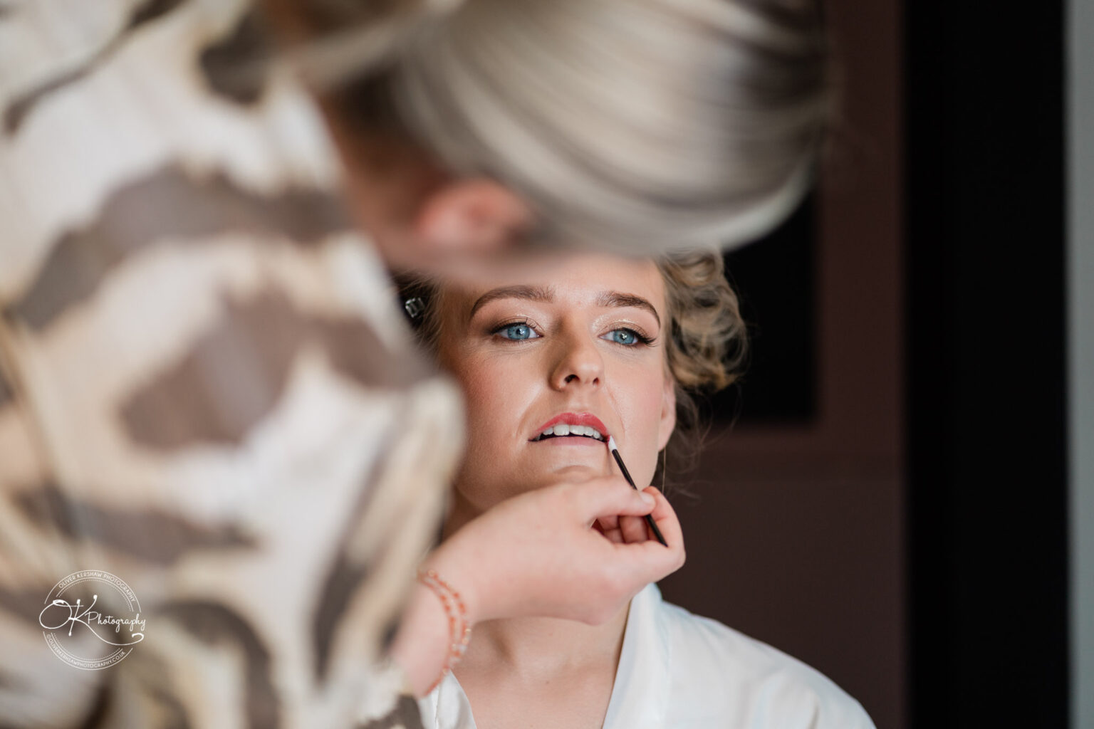 A close-up of a bride having lipstick applied, with a makeup artist's hand visible in the foreground.