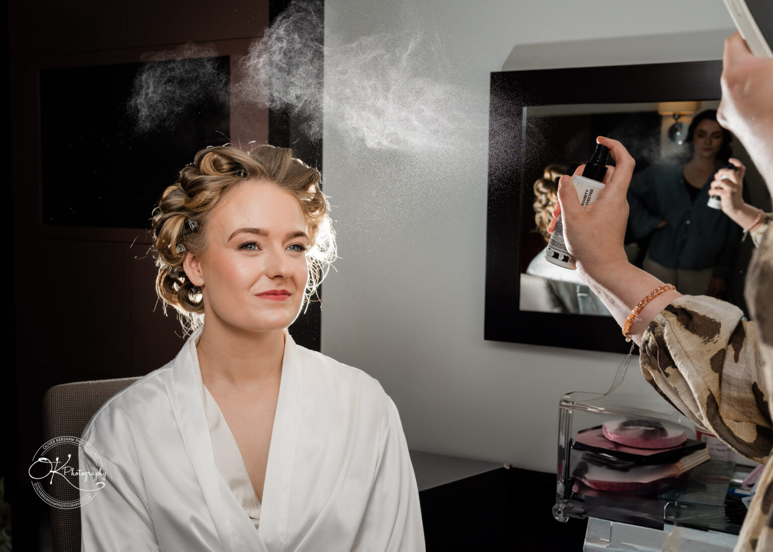 A bride having her hair styled, with a mist of hairspray visible in the air, as she sits calmly in a dressing gown.