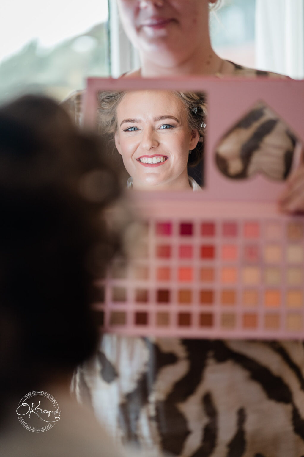A reflection of a bride smiling in a makeup palette mirror, with the makeup artist faintly visible.
