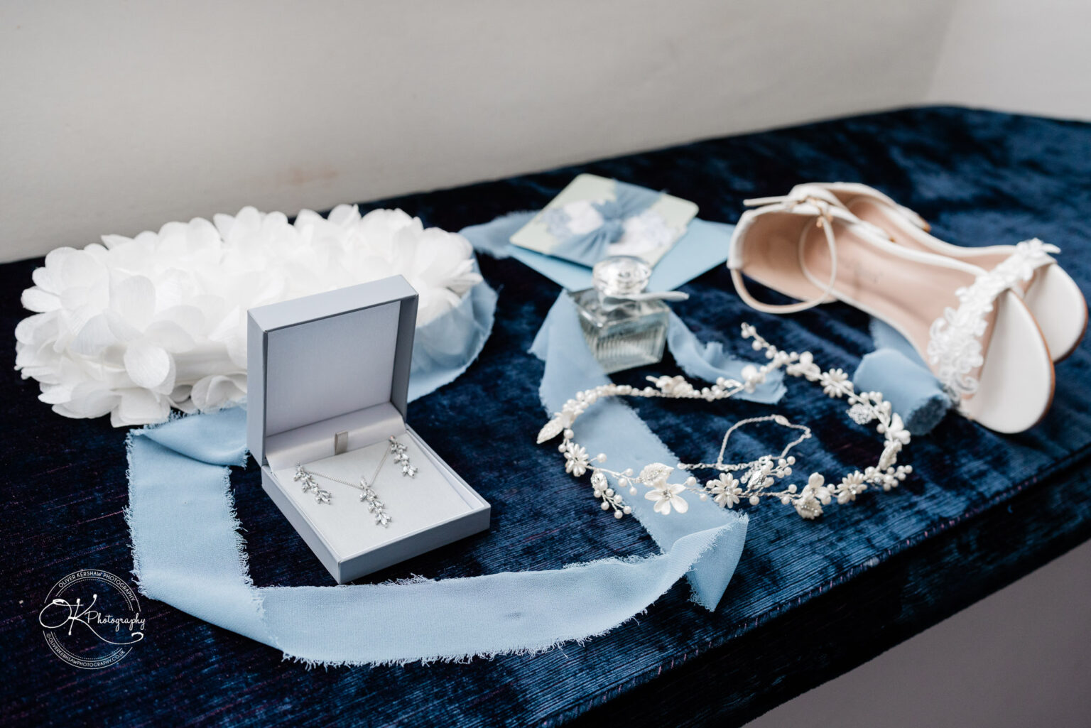 A flat lay of wedding accessories, including a necklace, earrings, perfume, a flower crown, and bridal shoes.