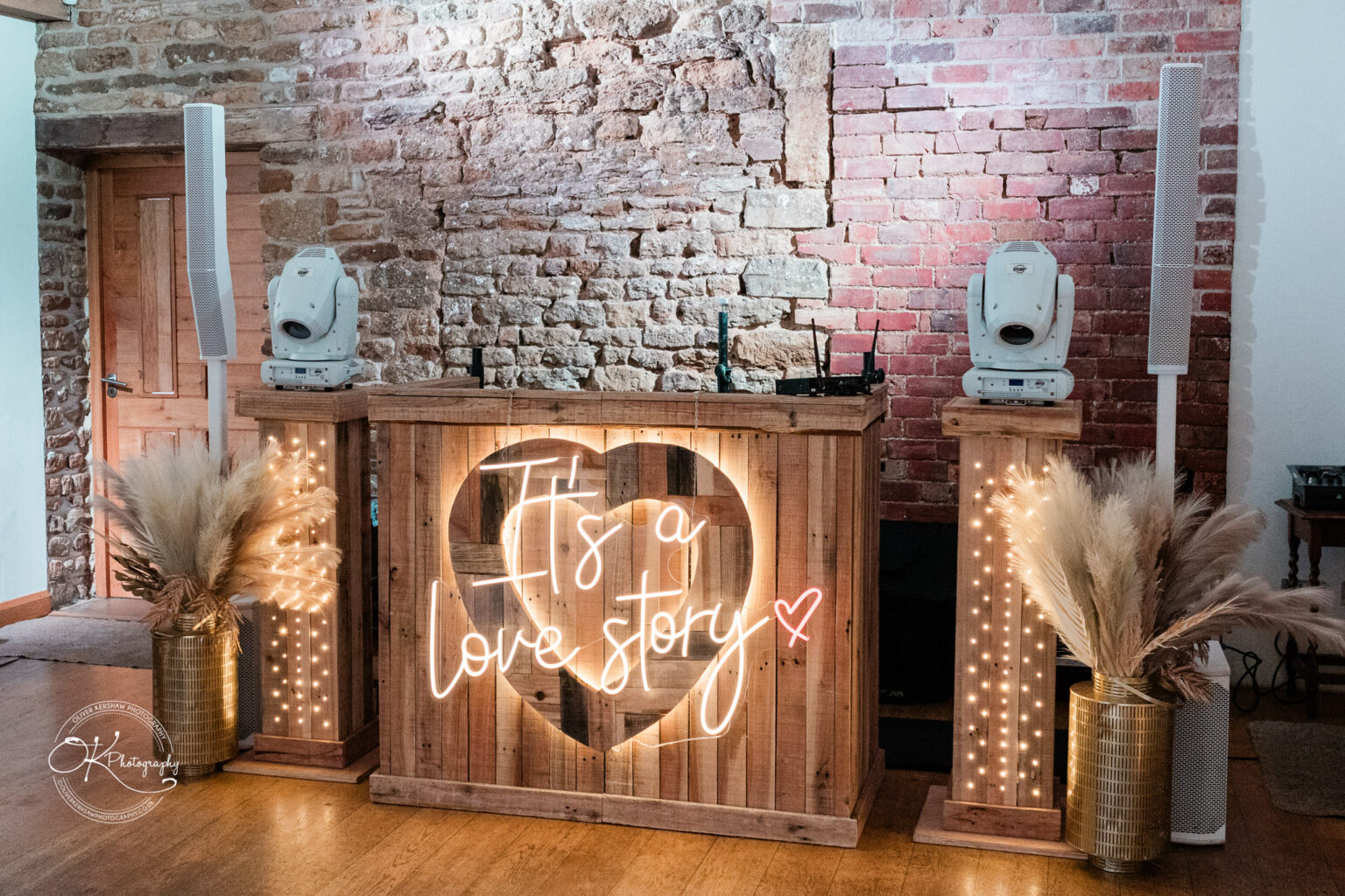 A wooden DJ booth with a neon sign reading 'It's a love story,' set against a rustic brick wall.