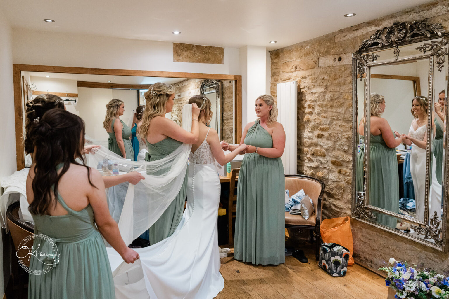 Bridesmaids helping the bride with her veil and gown in a cozy dressing room with a large mirror.