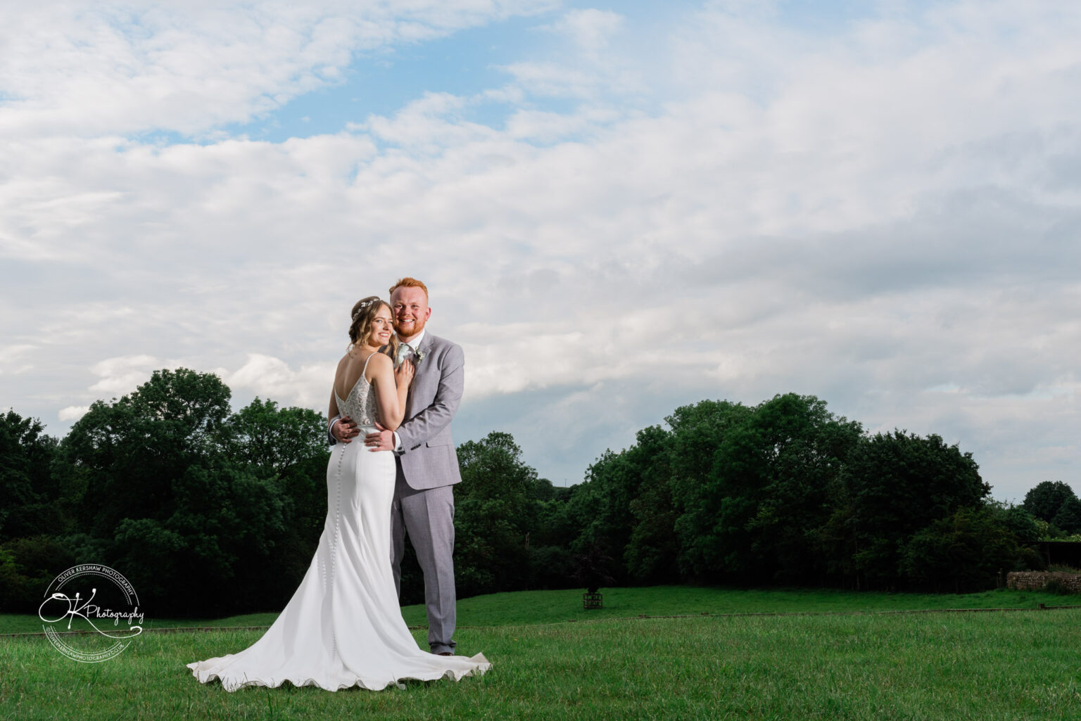 The bride and groom stand closely together, posing in a green field under a blue sky at Dodford Manor.