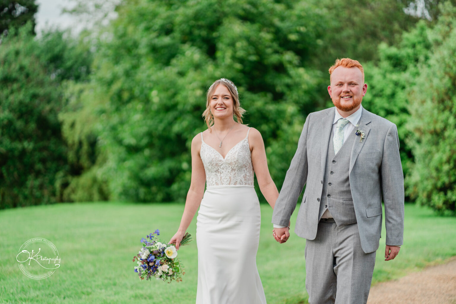 The bride and groom holding hands and smiling as they walk through a lush green area at Dodford Manor.